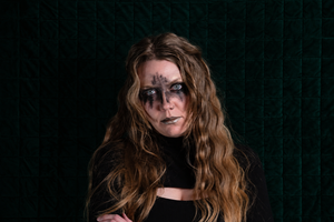 A woman with long wavy light brown hair and dramatic black face paint around her eyes and mouth, wearing a black top, posed against a dark green quilted background.
