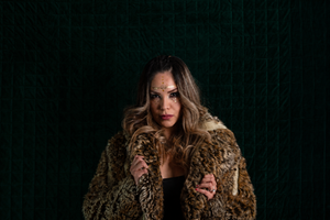 A woman with long wavy brown hair and a black top models a brown and beige shaggy faux fur coat against a dark green textured backdrop.