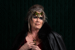 A person with gray hair styled in an updo, wearing a gold crown with green leaves, a black fur-trimmed coat, and a red top, holding a small gold and black pendant against a dark green textured background.
