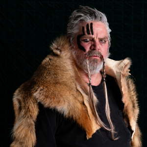 A man with gray hair and a beard wearing a brown fur collar over a black shirt, with black face paint on his forehead, photographed against a dark textured background.