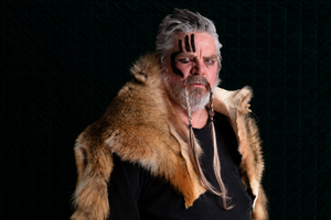 A man with gray hair and a beard wearing a brown fur collar over a black shirt, with black face paint on his forehead, photographed against a dark textured background.