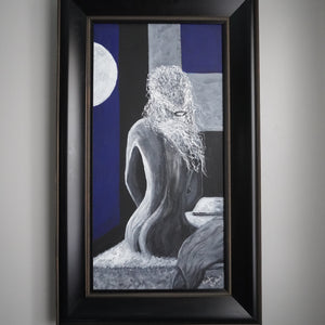 A framed surreal art print featuring a gray stone-like seated figure with long hair against a blue and gray background, enclosed in a black frame and hung on a light gray wall.